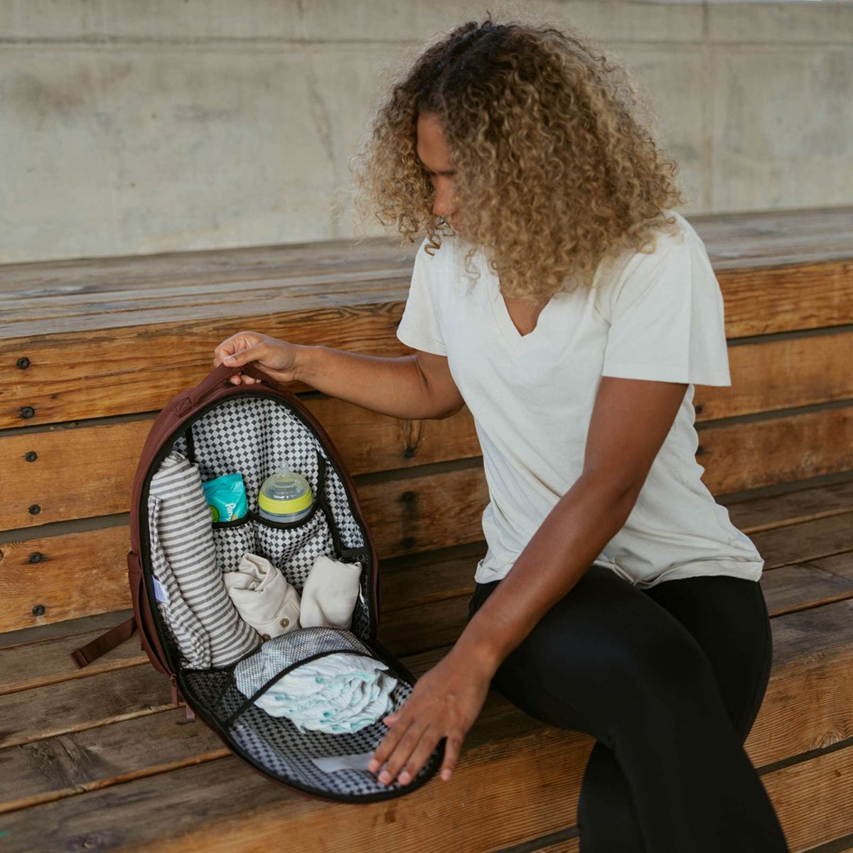 Woman sitting on a wooden bench with a backpack containing baby items.