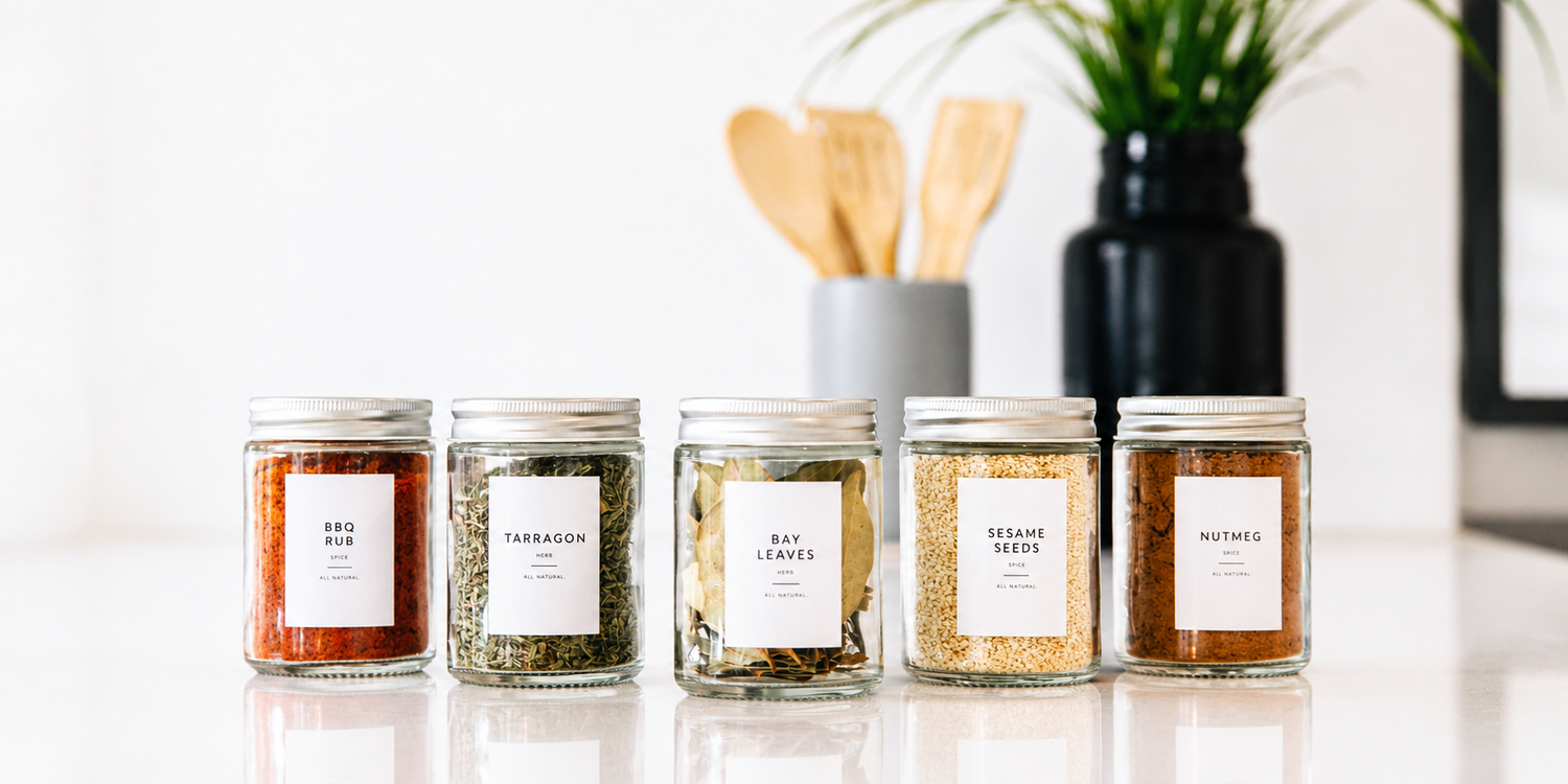 Row of clear glass spice jars with silver lids and minimalist white labels on a bright white marble countertop