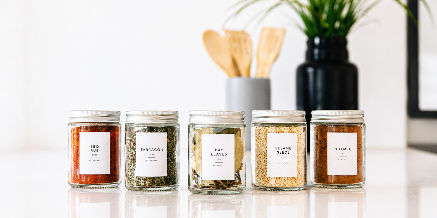 Row of clear glass spice jars with silver lids and minimalist white labels on a bright white marble countertop