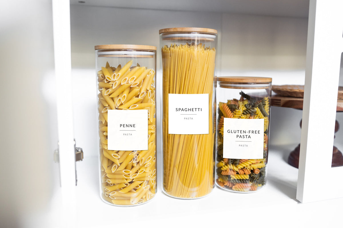 Labeled glass pasta jars on a white kitchen cabinet shelf