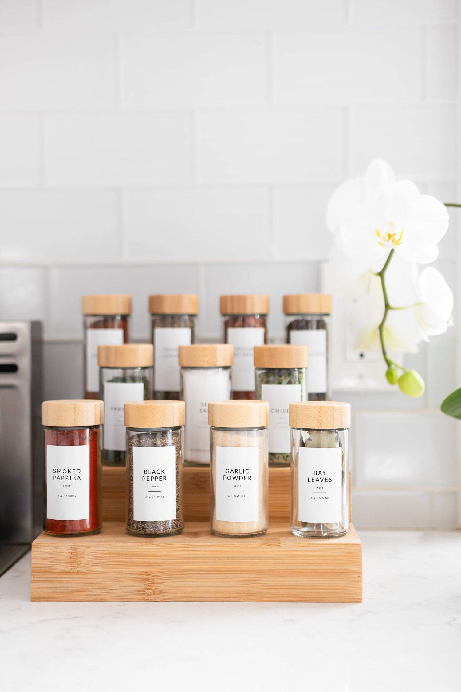 Tiered bamboo spice rack with glass jars and white minimalist spice labels on white marble counter - Savvy & Sorted