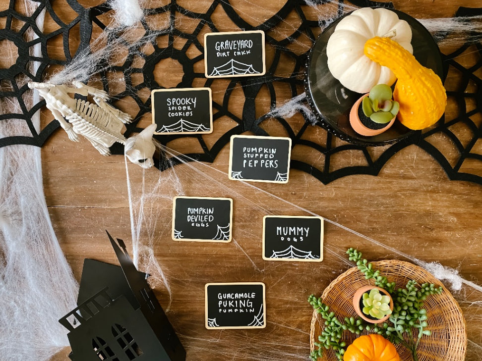Halloween party food spread labeled with mini chalkboard signs including Graveyard Dirt Cake, Spooky Spider Cookies, and more