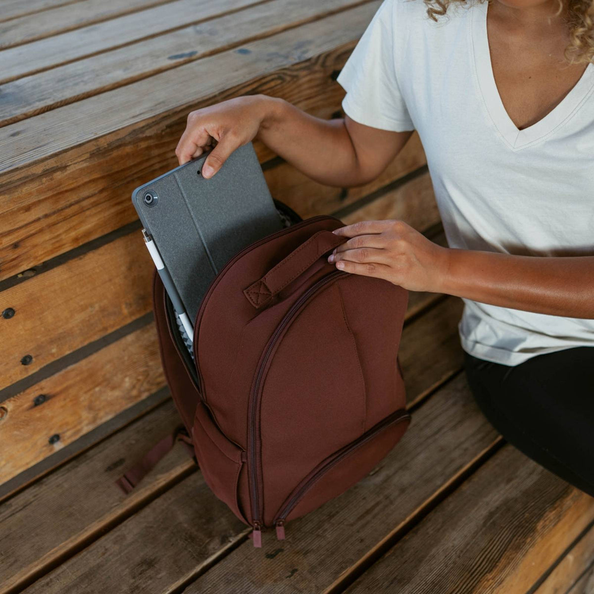 Person placing a tablet into a brown backpack on a wooden surface