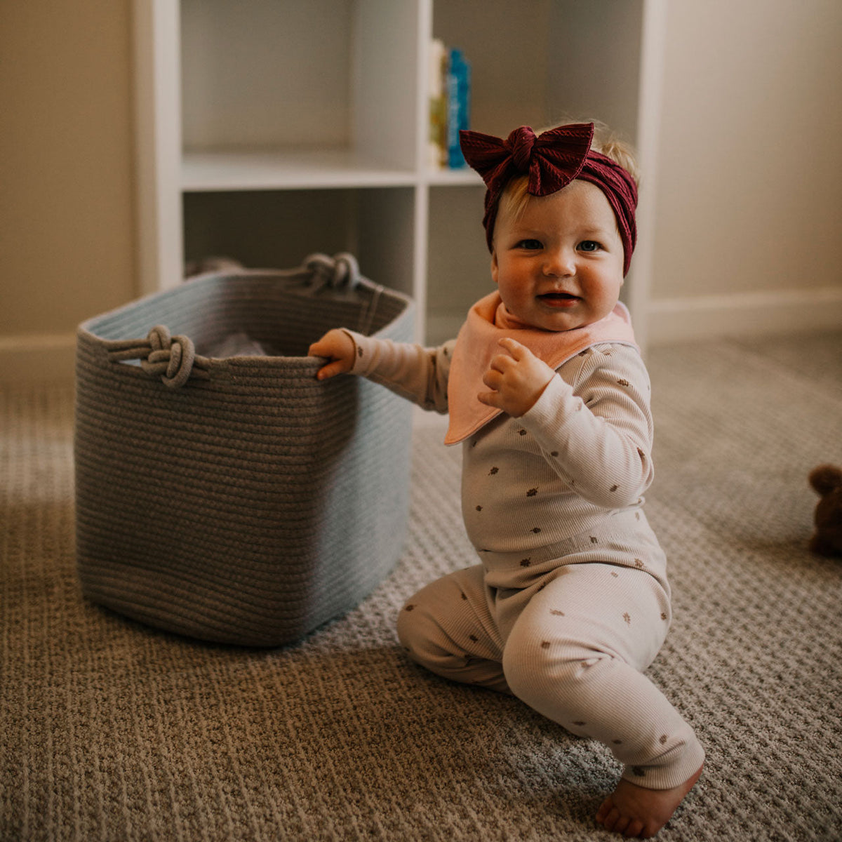 Baby in a polka dot outfit and headband sitting on the floor next to a woven basket.
