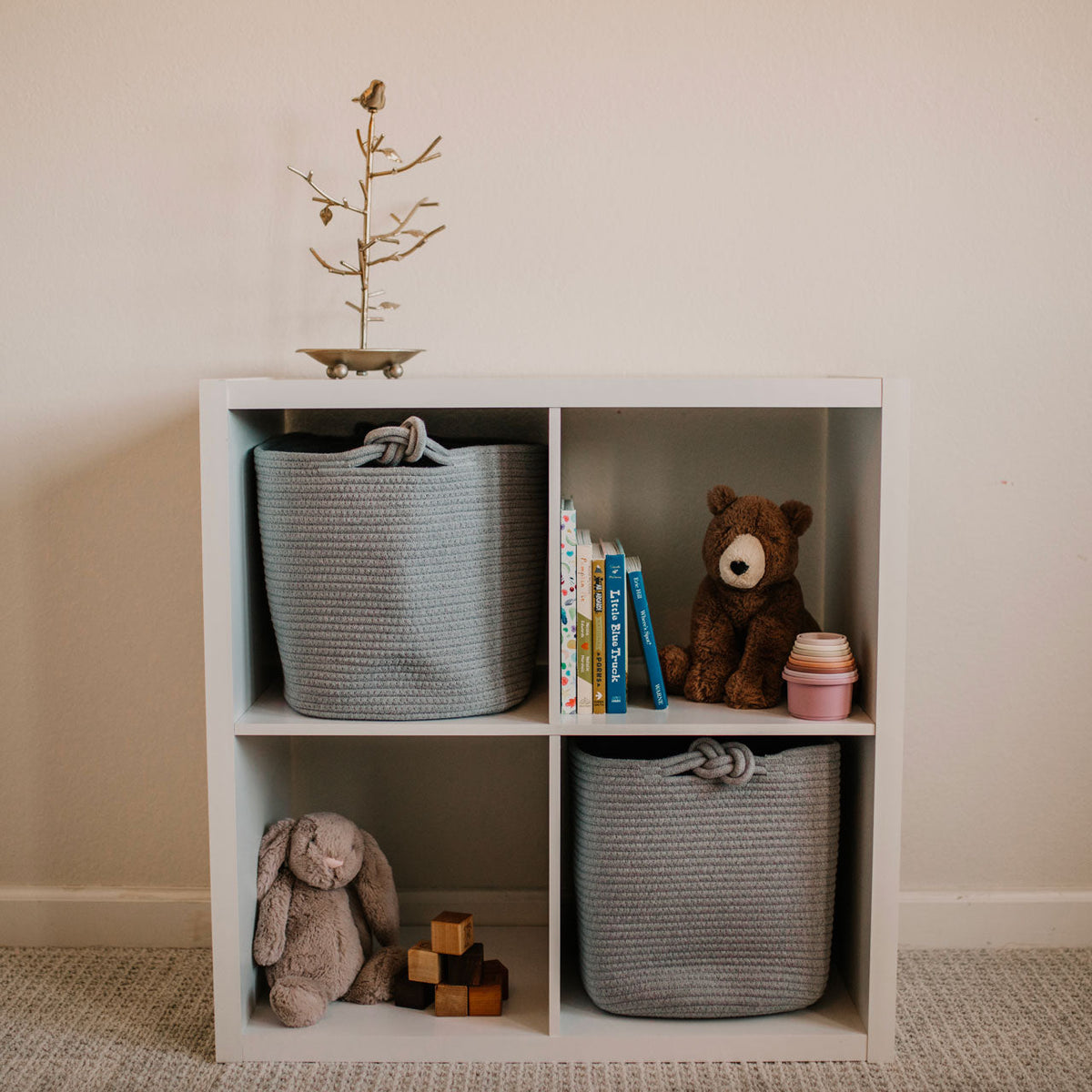 White cube shelf with storage baskets, books, and toys against a beige wall.