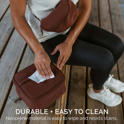 A person sitting on wooden deck boards uses a wipe to clean a brown neoprene pouch bag. The image features a matching brown crossbody bag worn by the person and text overlay at the bottom that reads &