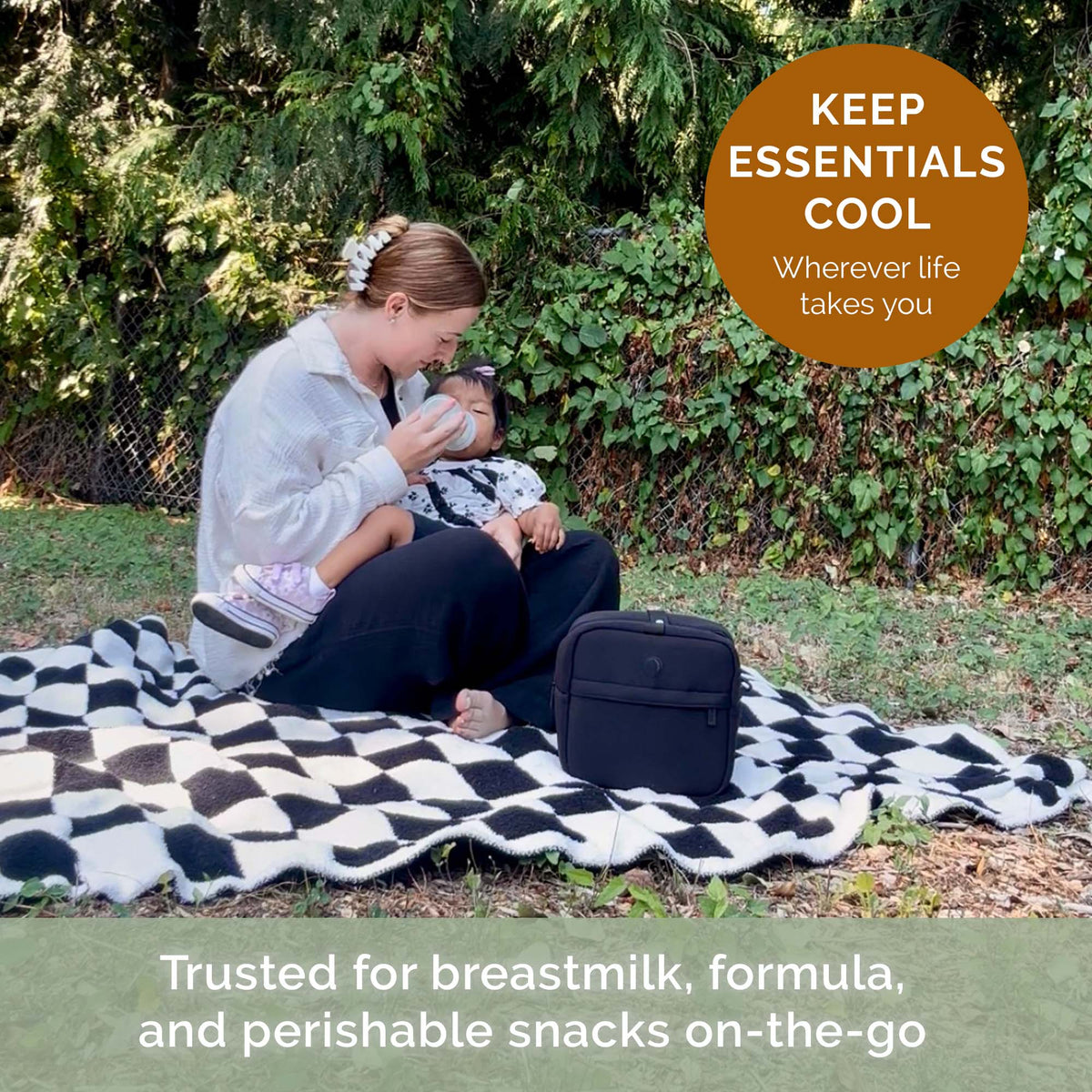 A woman and child sit on a black-and-white patterned blanket outdoors in front of a leafy green hedge, with a black insulated bag beside them. Text overlays read &