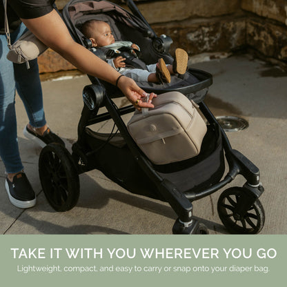 A beige diaper bag with a front zip pocket is shown attached to a black stroller in an outdoor setting, with a baby sitting inside and a person reaching toward the bag. A text banner at the bottom reads &