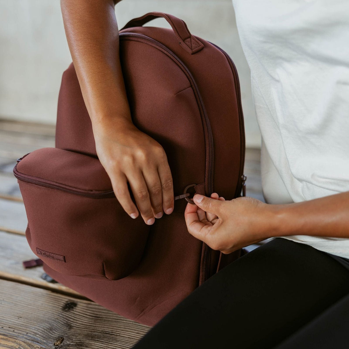 Person opening a brown backpack on a wooden floor.