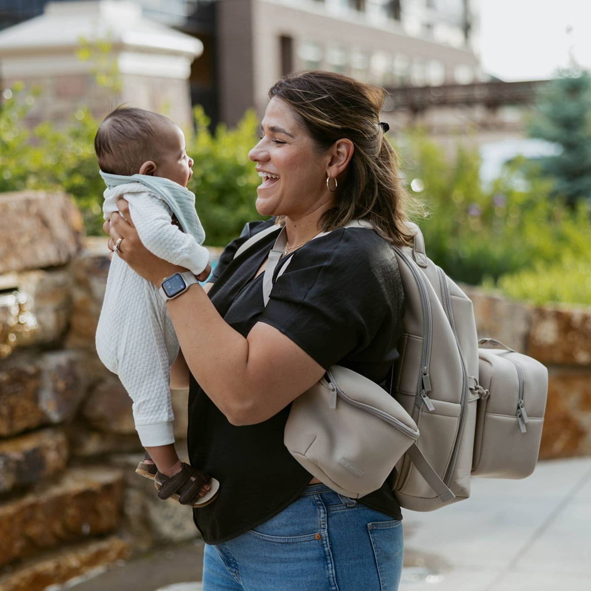 Woman holding a baby and wearing a beige backpack outdoors