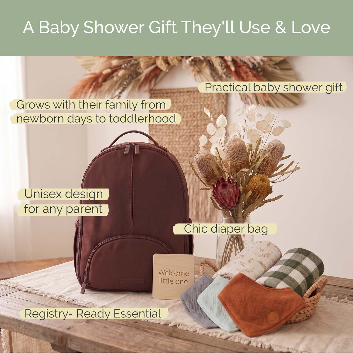 A brown diaper backpack displayed on a table with baby accessories and overlaid marketing text. The scene includes a basket of folded blankets, a vase of dried flowers, and a small welcome card, with labels such as &